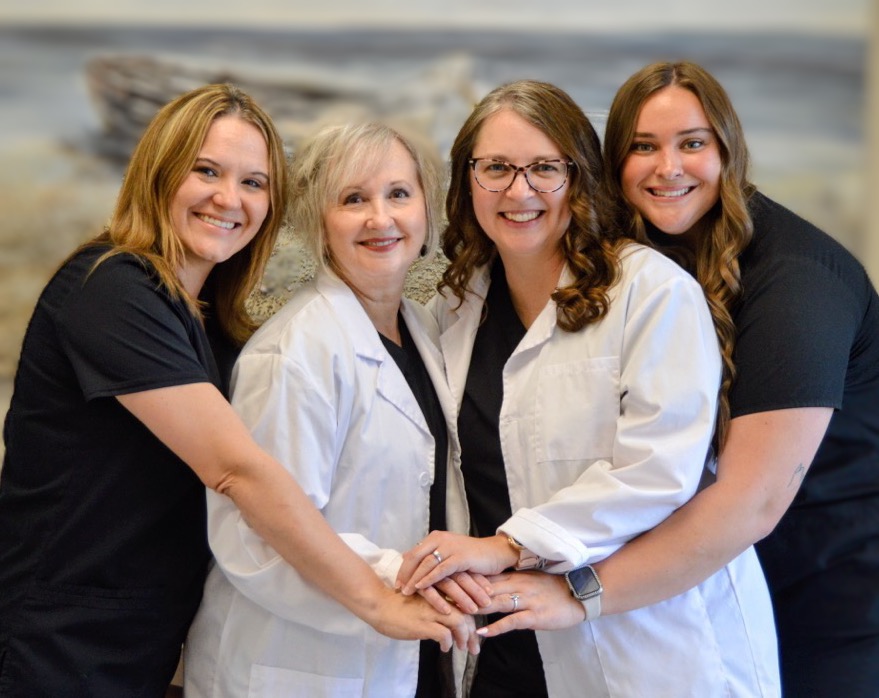 Four women smiling and holding hands, two in lab coats and two in black shirts.